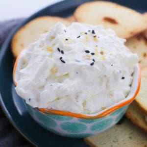 A bowl of creamy dip with everything seasoning on a plate with bagel chips