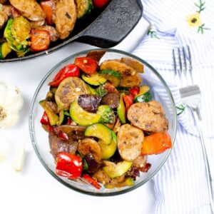 A cast iron skillet with sausage and veggies and a bowl in front of the skillet with a serving of the sausages. A fork is to the right of the bowl.