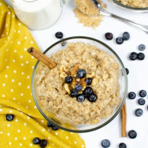 A glass bowl with cooked oatmeal, blueberries, nuts, and cinnamon stick.