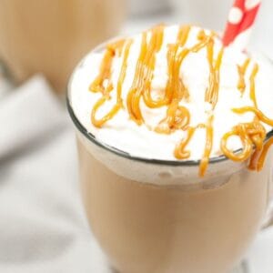 A clear mug filled with latte and topped with whipped cream and drizzled with caramel.