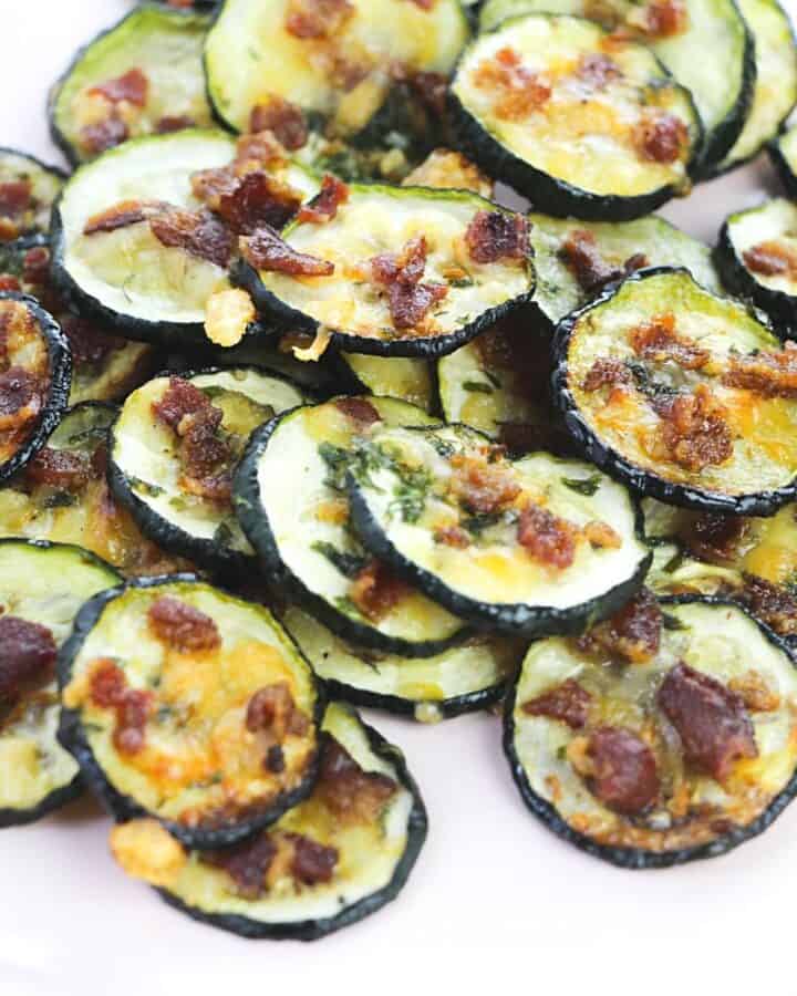 A pile of baked zucchini slices topped with bacon and cheese on a white plate.