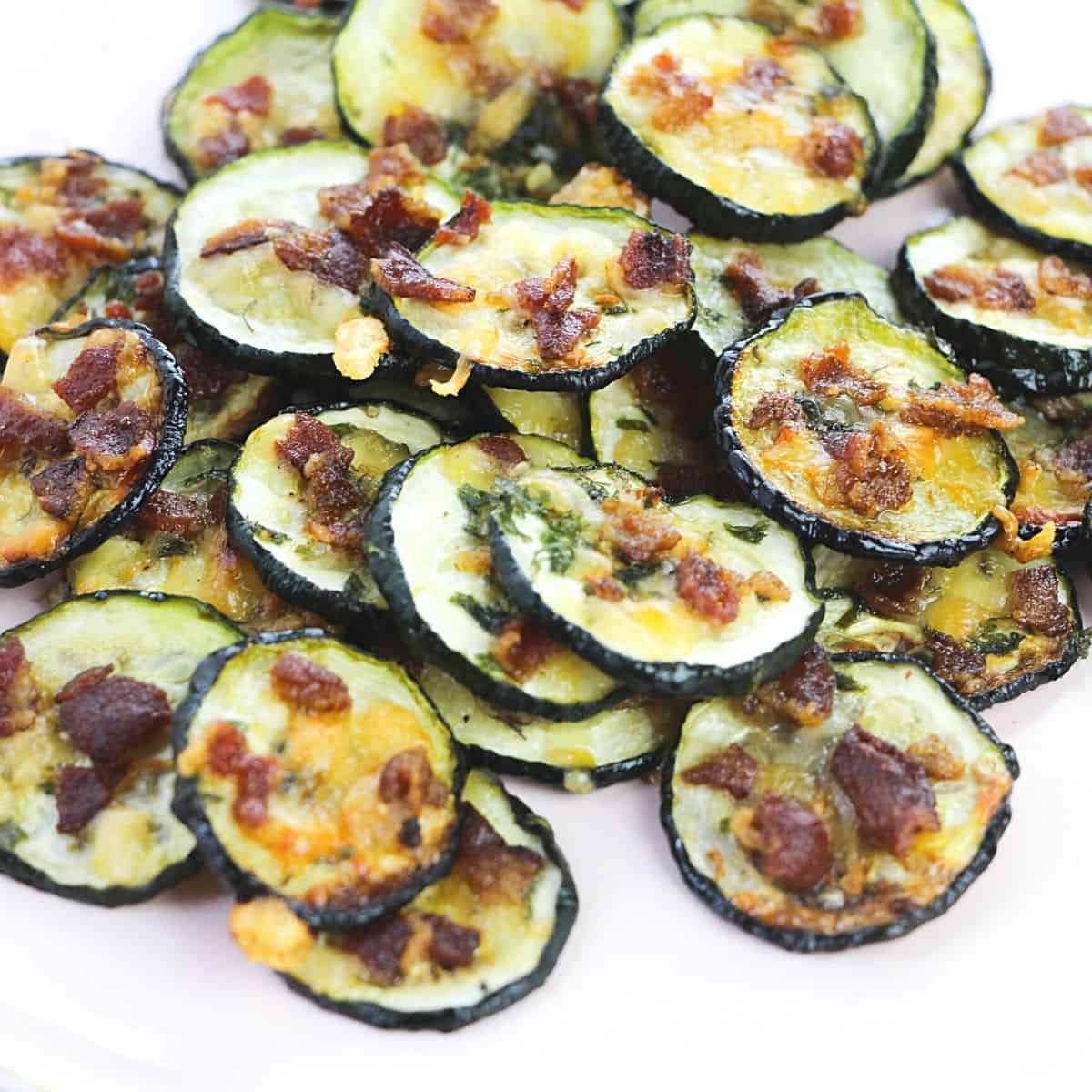 A pile of baked zucchini slices topped with bacon and cheese on a white plate.