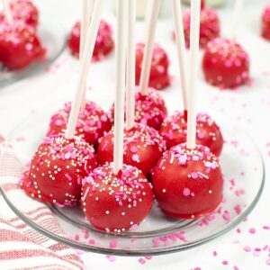 Cake pops with a red coating and white and pink sprinkles on a glass plate.