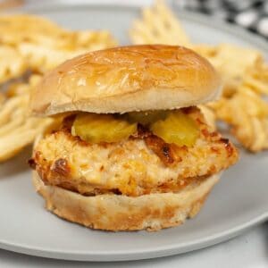 A chicken sandwich topped with pickles with waffles fries on a grey plate.