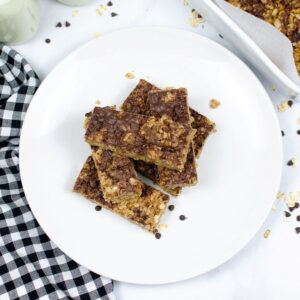 Homemade granola bars stacked on a white plate with a checkerboard napkin next to the plate.