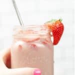 A hand lifting a glass mason jar filled with pink creamy drink, silver straw, and a strawberry on the side of the glass.