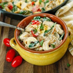 A yellow bowl with creamy tortellini, grape tomatoes, and fresh spinach in it.