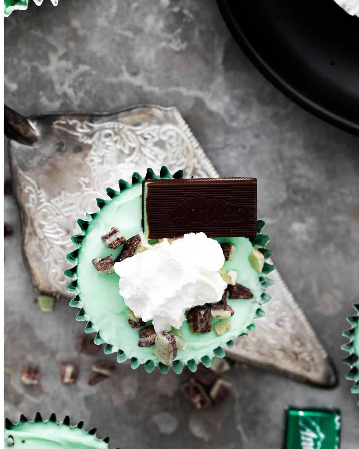 An overhead view of a silver serving spoon with a grasshopper mini cupcake with an Andes mint and whip cream on top of the cheesecake.