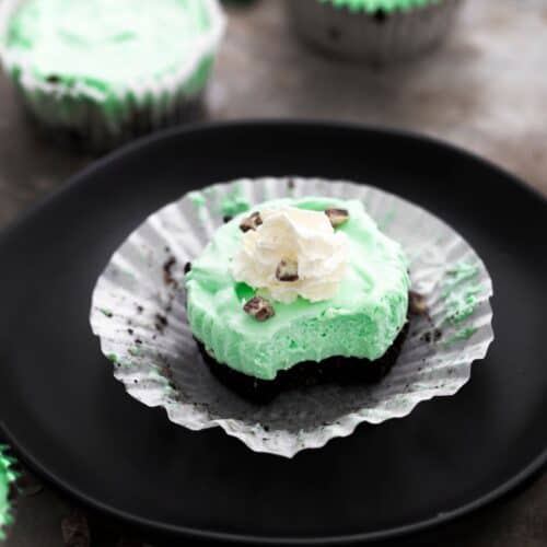 A mini cheesecake with an Oreo bottom and green tinted cream cheese topped with whip cream and chopped mints on an open cupcake liner.