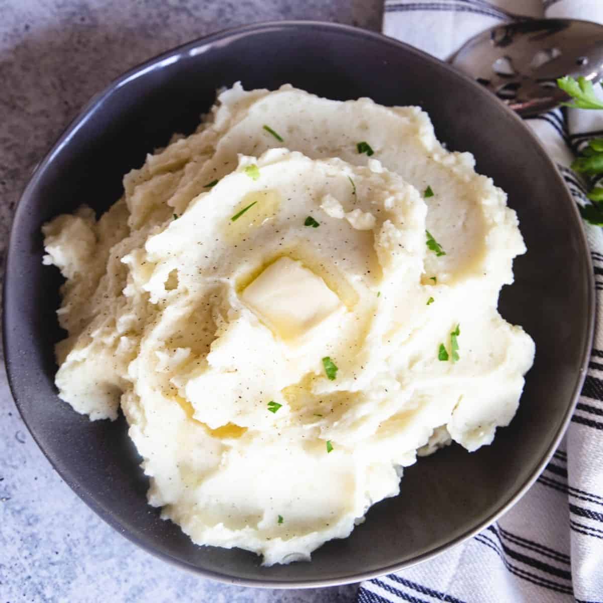 A grey round bowl with homemade mashed potatoes topped with chopped parsley and melting butter.