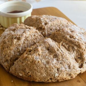 A round loaf of brown bread on a wood board with a yellow bowl of strawberry jam behind the brea.