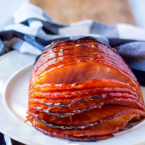 A sliced glazed ham on a plate with a blue and white checkered napkin behind it.