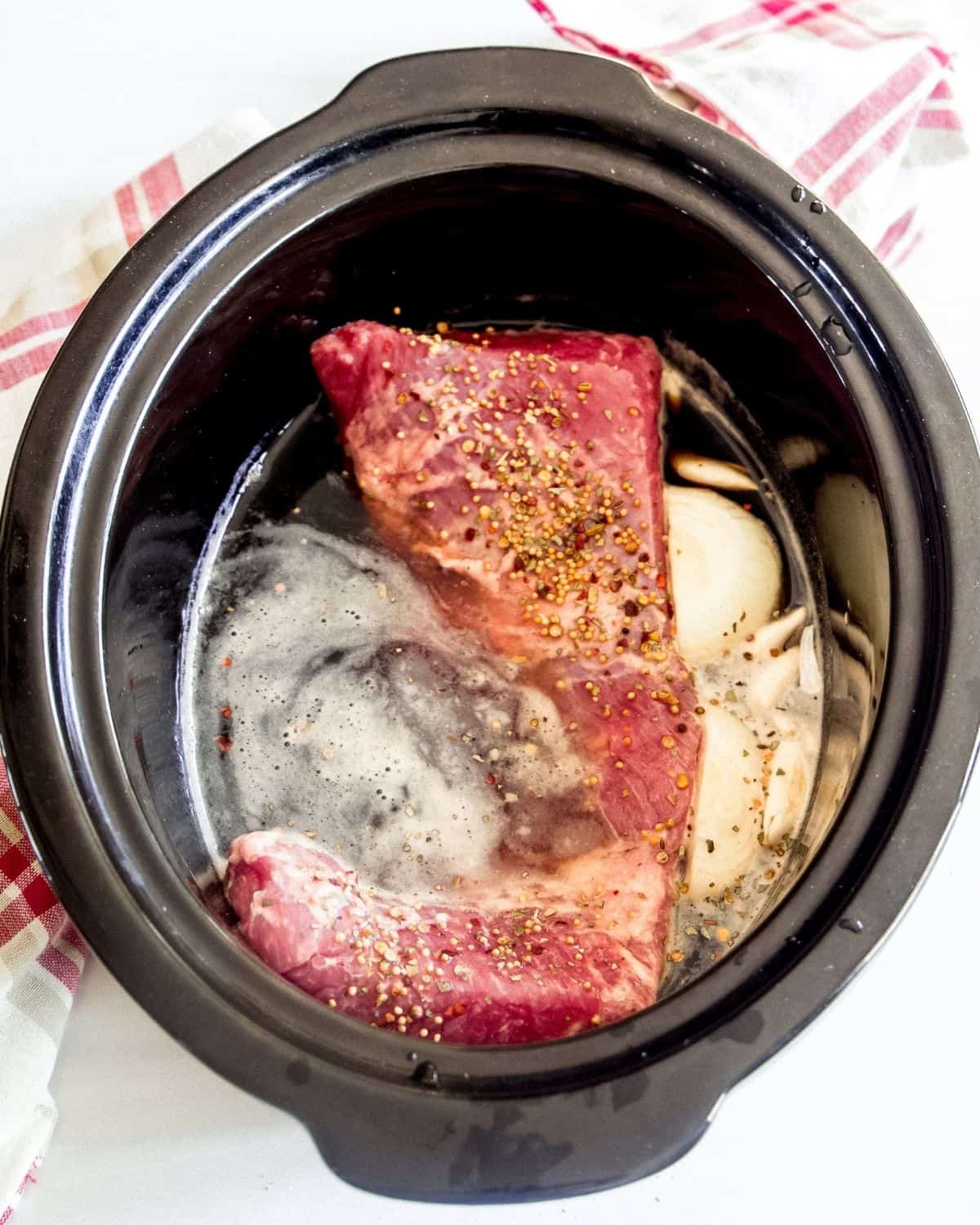 A black slow cooker vessel with a corned beef in the pot. The beef is in a Guinness beer broth and pickling spices.