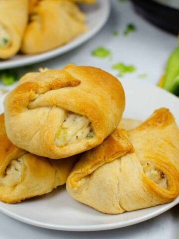 A trio of chicken stuffed crescent rolls on a plate with another plate behind it filled with chicken sandwiches.