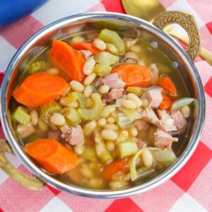 A silver bowl with gold handles filled with soup, carrots, celery, beans, and diced ham.