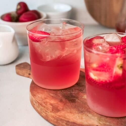 Two glasses of strawberry iced tea, ice, and sliced strawberries sitting on a wooden tray.