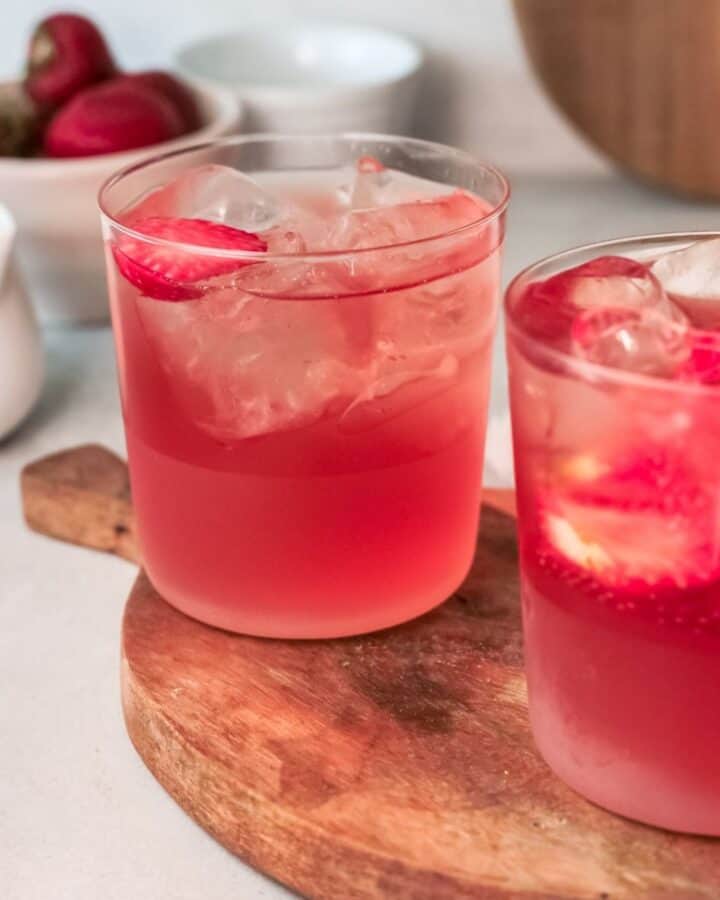 Two glasses of strawberry iced tea, ice, and sliced strawberries sitting on a wooden tray.