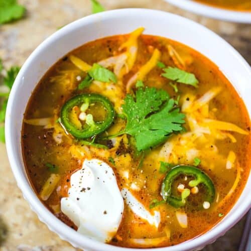 A white soup bowl fillled with enchilada soup topped with melted cheese and a dollop of sour cream.