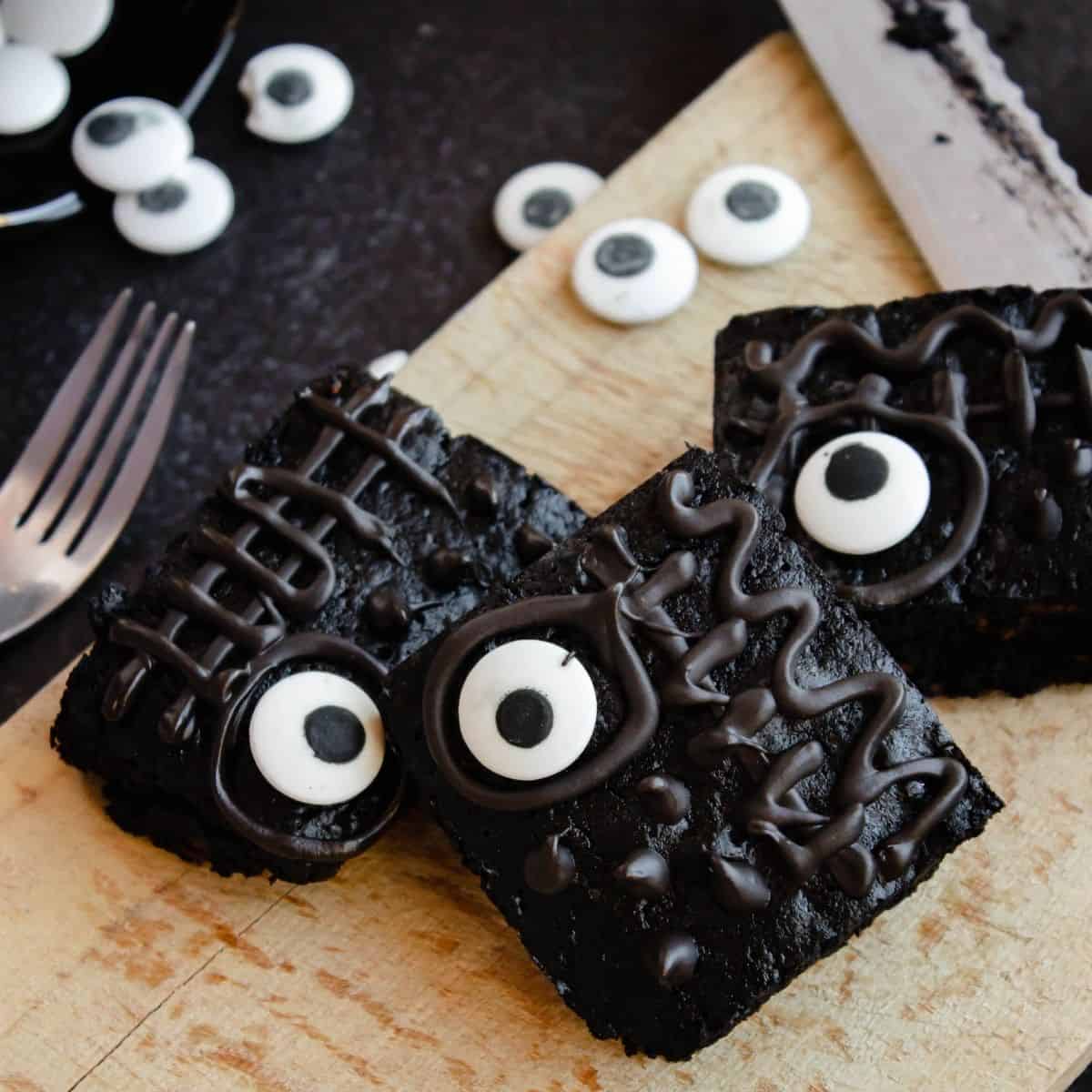 Three brownies with candy eye balls and chocolate frosting drizzled on the top of the brownies. A knife is next to the board.