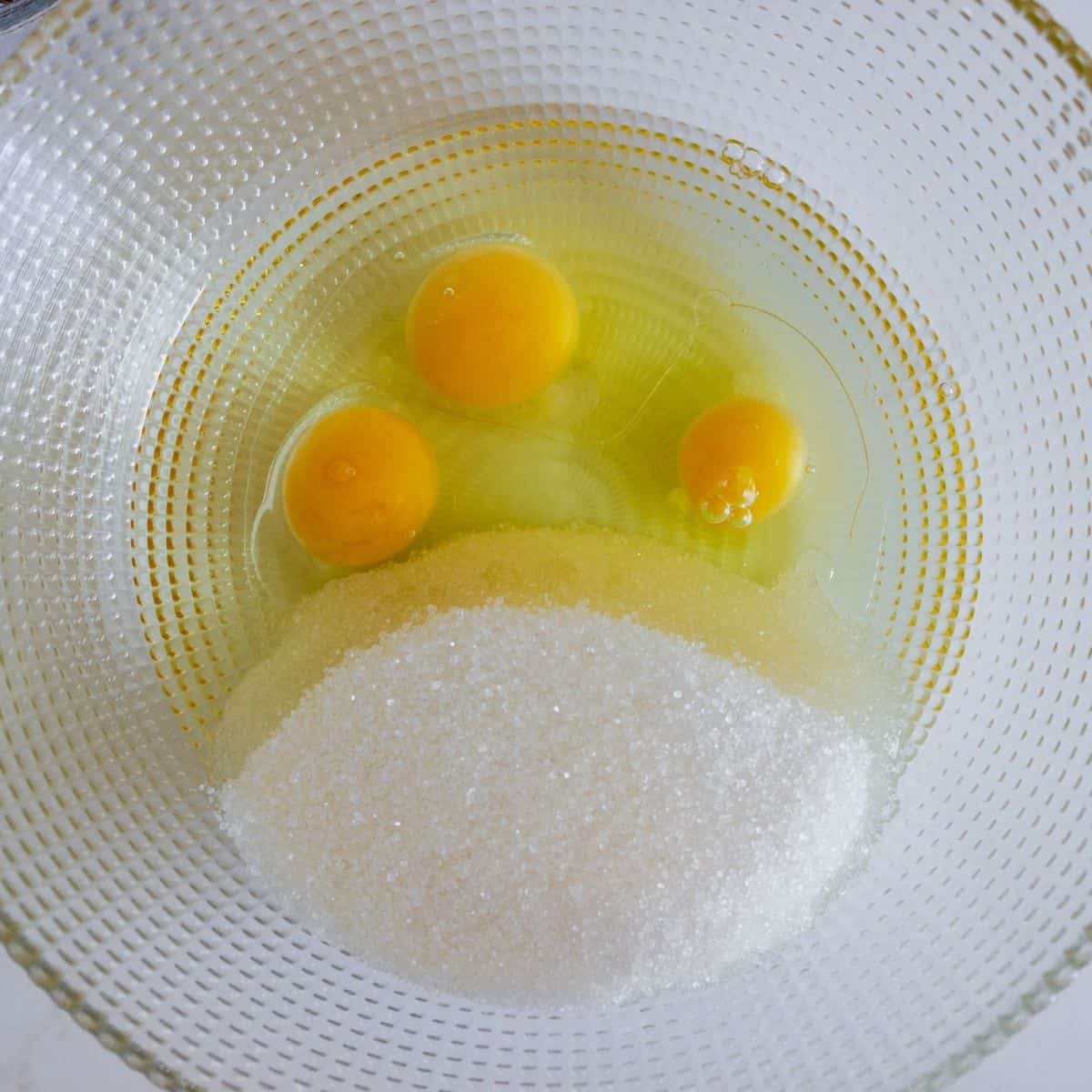 A glass bowl with 3 eggs out of the shell and sugar, not blended together.