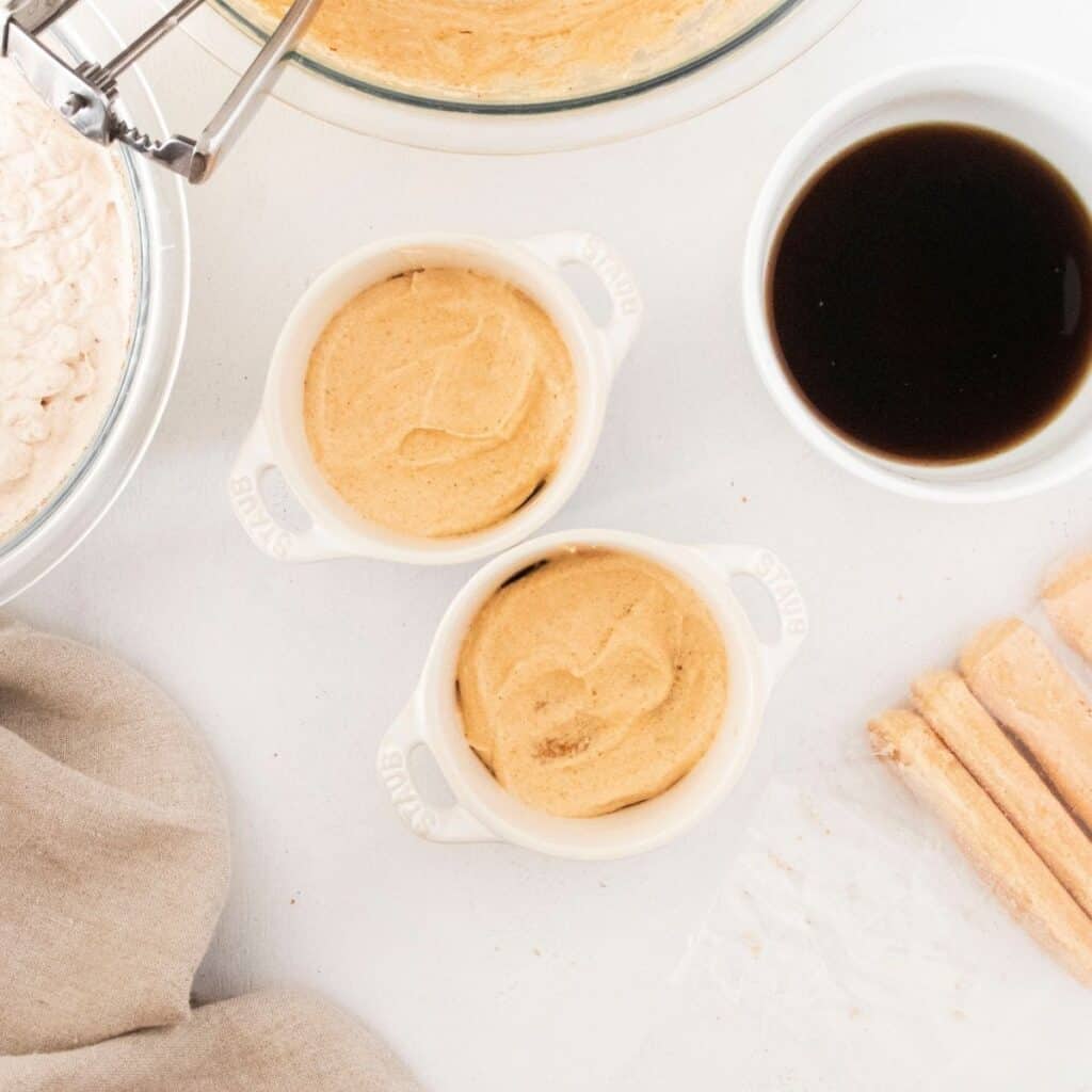 Two white cocottes cups with pumpkin mousse inside. Espresso in a bowl next to it and ladyfinger pastry on the side. 