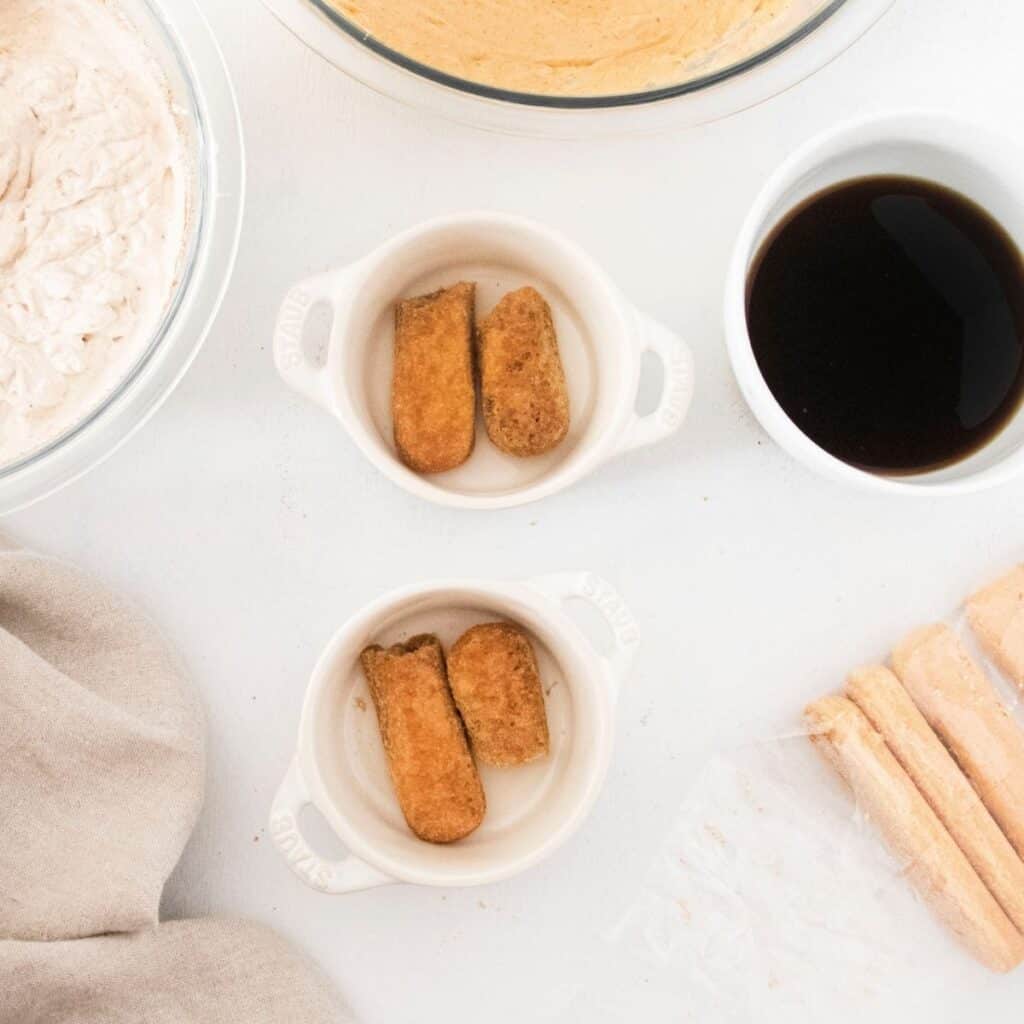 Lady fingers split in half in the bottom of a white ramekin and a bowl of espresso next to the ramekins.