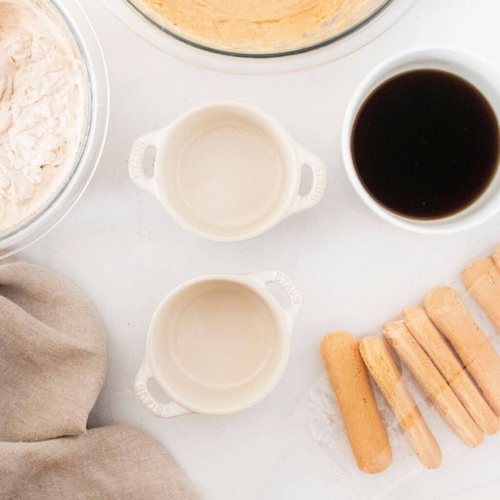 Two small ramekins and a bowl of dark liquid and lady fingers on a white table. 