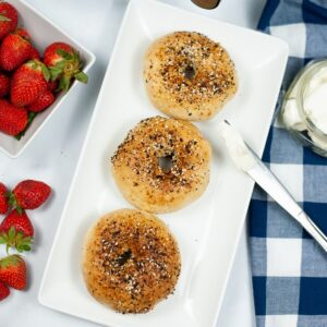 A white rectangle cake with a knife and cream cheese leaning on the plate. Three everything bagels on the plate with strawberries on the side.