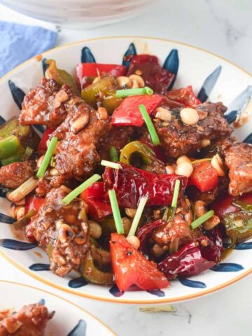 A white bowl on a blue linen with a blue painted lines filled with chicken with chicken in a brown Asian sauce.