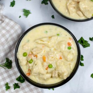 A bowl of chicken soup with dumplings and peas with chopped parsley on the side of the bowl.