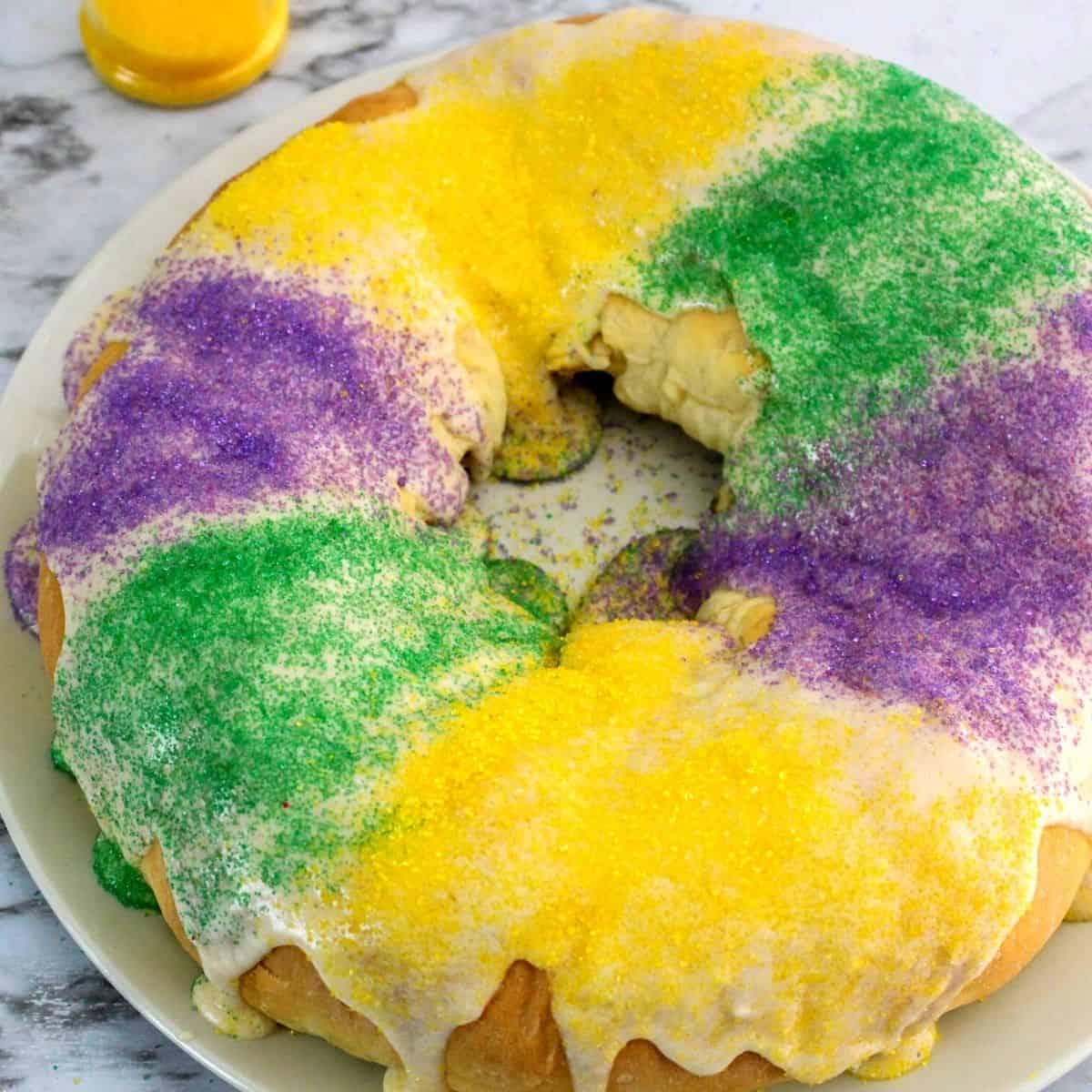 An overhead view of a round cake with white icing and purple, green, and yellow sprinkles.