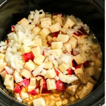 A black slow cooker vessel filled with chopped potatoes, corn, onions, and broth.