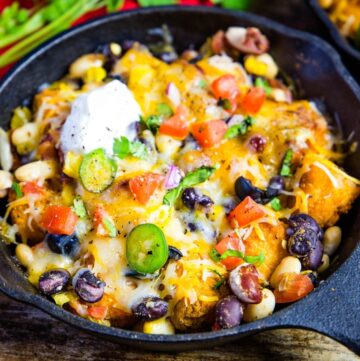 A cast iron skillet with cheese covering tater tots and beans sprinkled with chopped cilantro, jalapeno slices, and sour cream.