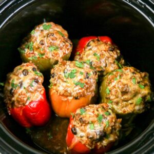 A slow cooker vessel with colorful peppers stuffed with beef and beans, topped with melted cheese.