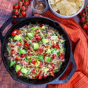 A cast iron skillet with ground beef topped with avocado, tomatoes, and cheese.