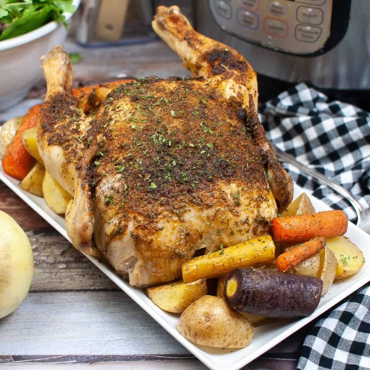A white platter with a golden brown roast chicken on the platter with roasted carrots, purple carrots and potatoes around the chicken.