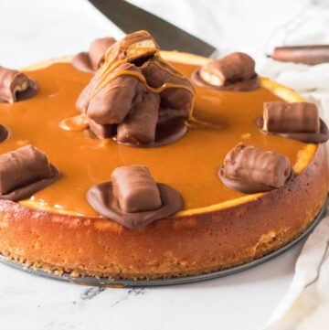 A Twix cheesecake topped with a brown sweet sauce, the center of the cake has 4 half pieces of a chocolate bar. The rim of the cake has chocolate rounds topped with Twix bars.