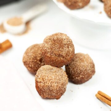 Four churro balls on a white plate with cinnamon sticks on the side.