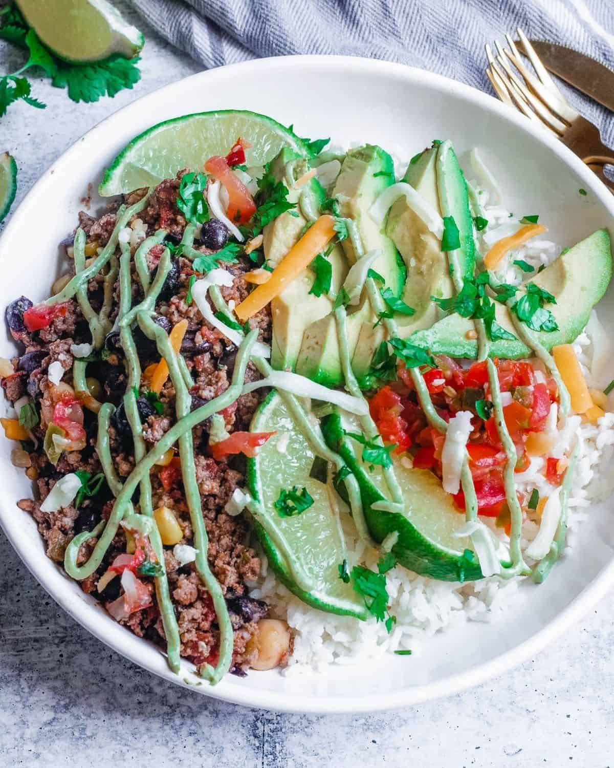 A white bowl with ground beef burrito topped with avocados, tomatoes, and avocado crema.