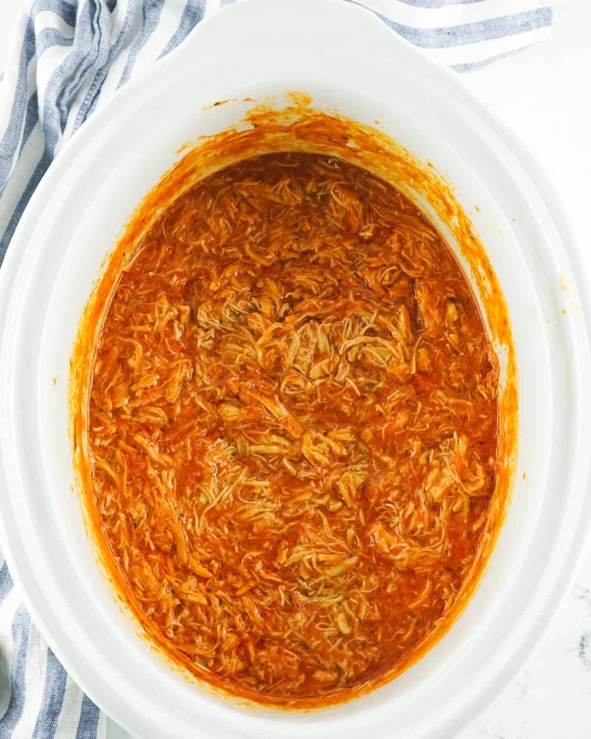 A slow cooker with shredded buffalo chicken to make shredded buffalo chicken sandwiches. 