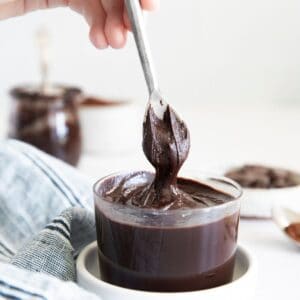 A glass bowl of hazelnut chocolate spread in a white bowl and a spoon pulling the chocolate out of the bowl.