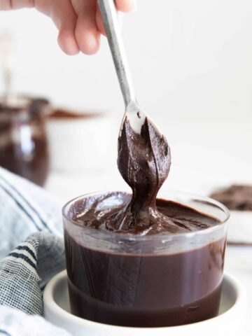 A glass bowl of hazelnut chocolate spread in a white bowl and a spoon pulling the chocolate out of the bowl.