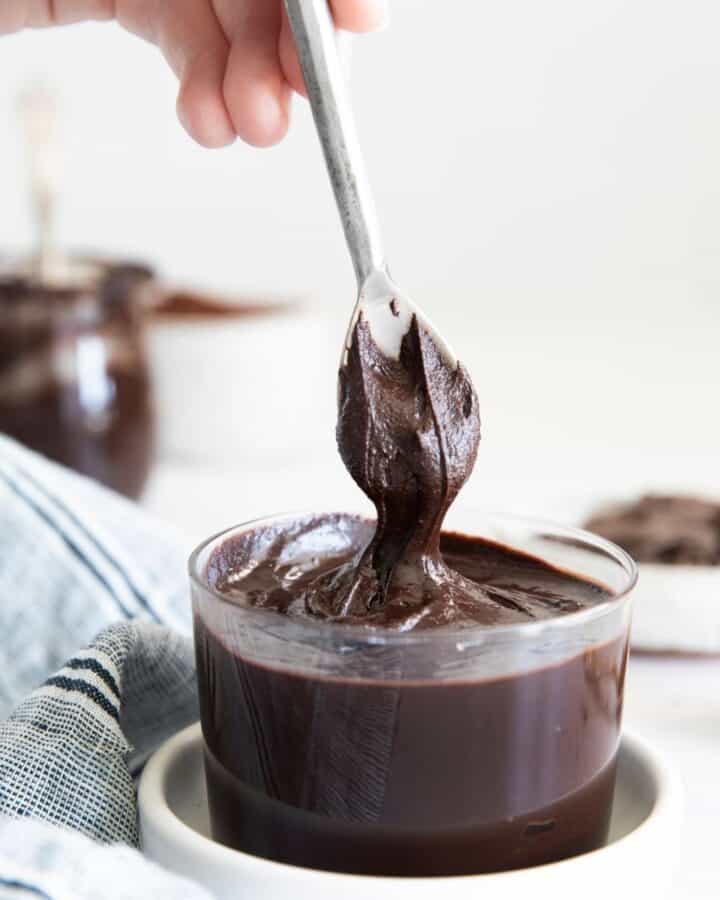 A glass bowl of hazelnut chocolate spread in a white bowl and a spoon pulling the chocolate out of the bowl.