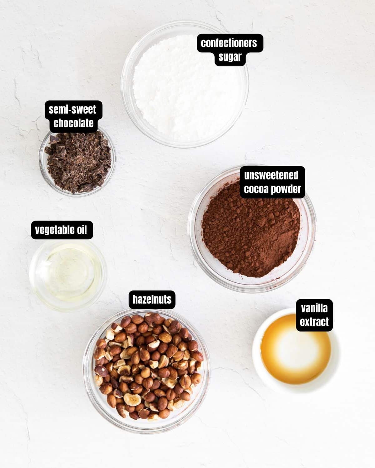 A photo with text overlay of ingredients; confectioners sugar, semi-sweet chocolate, unsweetened cocoa powder, vegetable oil, hazelnuts, vanilla extract on a white table.