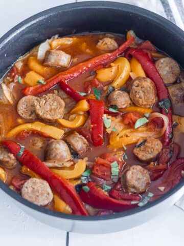 A saute pan filled with cooked sliced sausages, red peppers, yellow peppers in a red sauce.