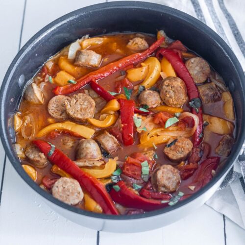 A saute pan filled with cooked sliced sausages, red peppers, yellow peppers in a red sauce.