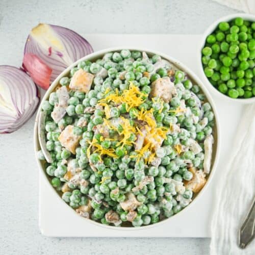 An overhead view of a white bowl filled with pea salad and bacon in a creamy white dressing topped with lemon zest.
