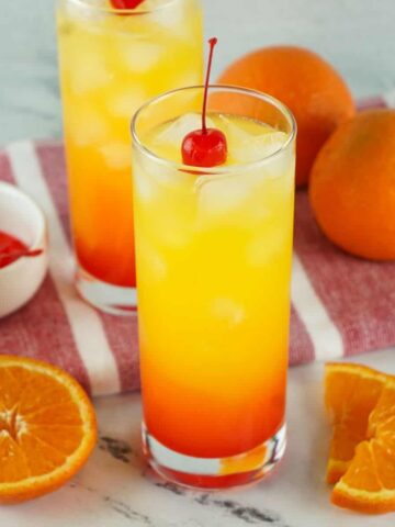 Two clear glasses with a red beverage at the bottom and a yellow liquid above the red, topped with a cherry.