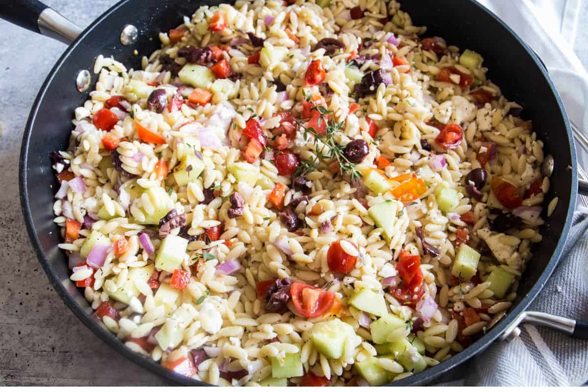 A large pan filled with the best orzo salad with chopped vegetables, sliced olives, and a sprig of thyme.