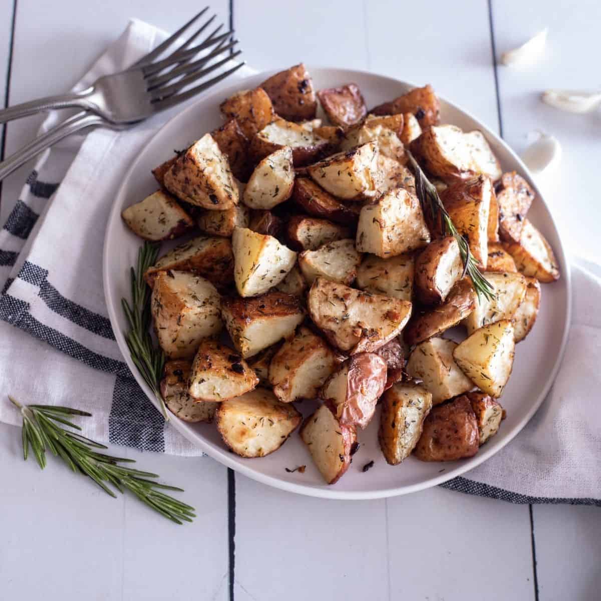 A white table with a large plate of roasted red potatoes on a white and black linen napkin under the plate.